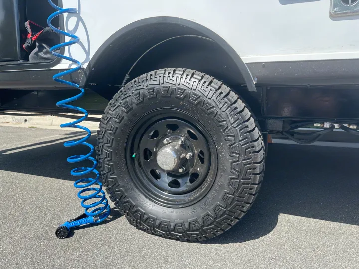 A rugged all-terrain tire on an RV rental vehicle, with a blue coiled hose and sprayer nearby for cleaning or maintenance.. Autumn Ridge 188BHS 2025