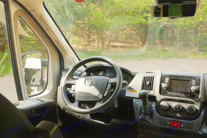 Dashboard and steering wheel of an RV rental, looking out the windshield at a wooded area.. Winnebago Solis 2022