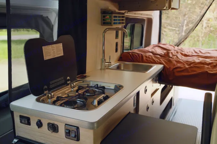 RV rental interior showcasing a compact kitchen with a two-burner stove, sink, and adjacent bed.. Winnebago Solis 2022