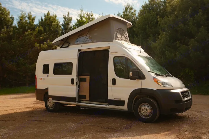 White camper van RV rental with pop-top roof extended, side door open, parked on a dirt road near pine trees.. Winnebago Solis 2022