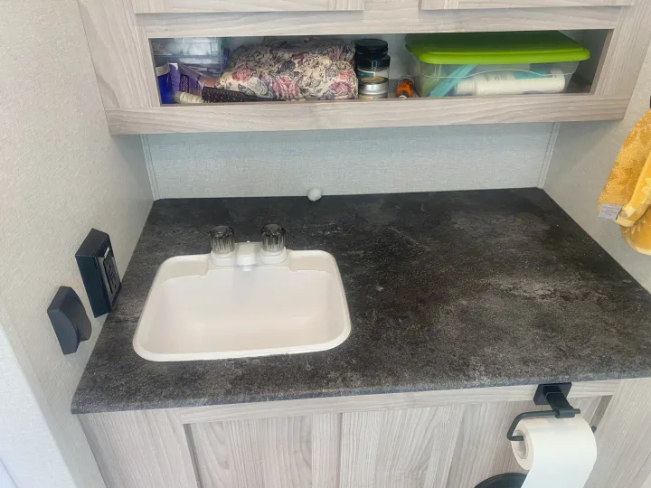 An RV rental bathroom, showing a compact sink, dark countertop, open shelves with toiletries, and a toilet paper holder.. Coachmen Northern  Spirit Ultra Lite 2024