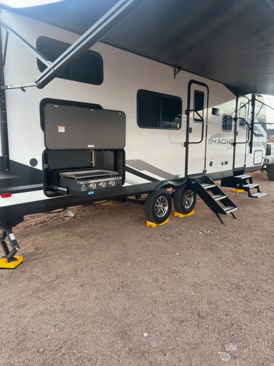 This white Imagine XLS RV rental features an extended awning and an open outdoor kitchen with a griddle, parked on gravel.. Grand Designs 25DBE 2025