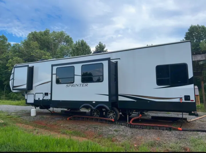 This white Sprinter travel trailer, an RV rental, is parked on gravel in a wooded area with slide-outs extended.. Keystone RV Sprinter Campfire Edition 2021