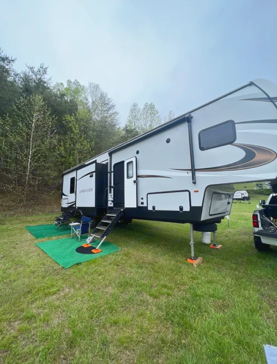 A large, modern fifth-wheel RV is parked at a grassy campsite with green mats & chairs, ready for your rv rental adventure.. Keystone RV Sprinter Campfire Edition 2021