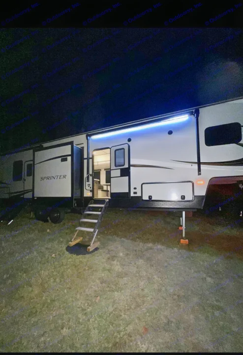 Night scene of a modern fifth wheel RV rental with interior and blue exterior lights on, steps down to grass.. Keystone RV Sprinter Campfire Edition 2021