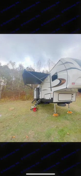 Fifth-wheel RV rental parked on grass with awning out, showing steps and leveling jacks, in a wooded outdoor setting under cloudy skies.. Keystone RV Sprinter Campfire Edition 2021