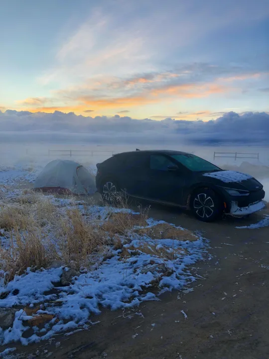 Winter camping: A car and tent are parked in a snowy, misty landscape with a fence. For next time, consider an RV rental!. Kia EV6 Wind All Wheel Drive 2022