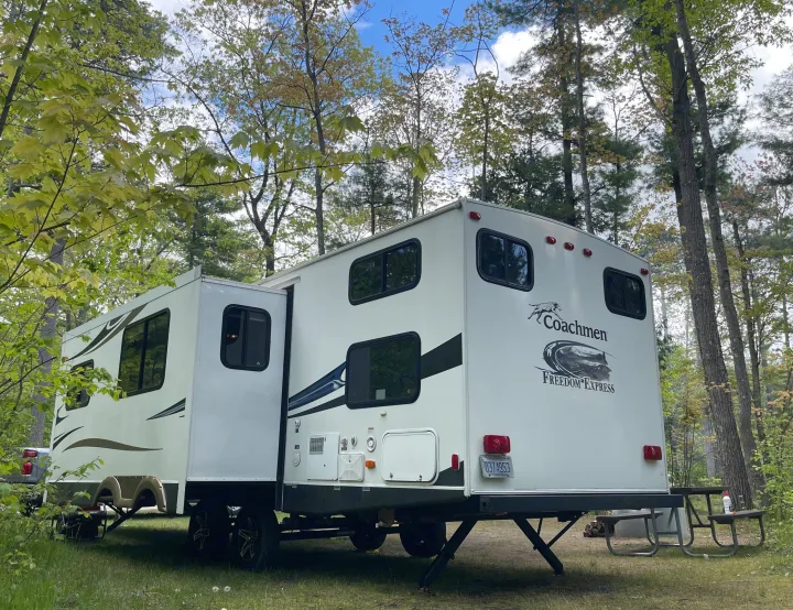 A white Coachmen RV rental is parked at a serene Michigan campsite, surrounded by trees. Shows its extended slide-out and picnic table.. Coachmen Freedom Express 2013