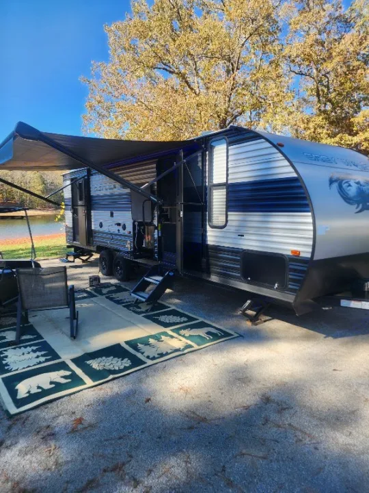 This RV rental travel trailer is set up at a beautiful fall lakeside campsite with an extended awning and an outdoor rug.. Forest River Cherokee Grey Wolf 2022