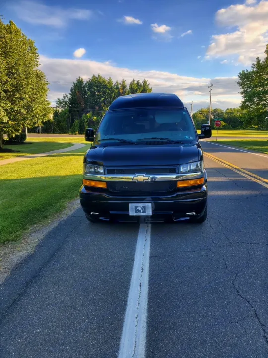 Front view of a sleek black high-top conversion van, ideal as an RV rental, parked on a road with a center line.. Chevy Luxury Splinter Express Express Conversion Van 2024