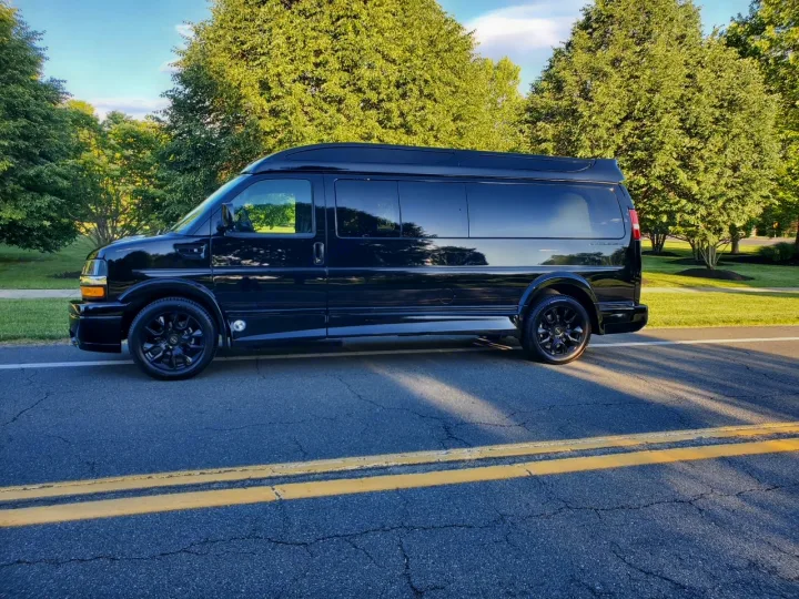 Sleek black custom Explorer conversion van for RV rental, parked on a road with double yellow lines, lush trees in background.. Chevy Luxury Splinter Express Express Conversion Van 2024