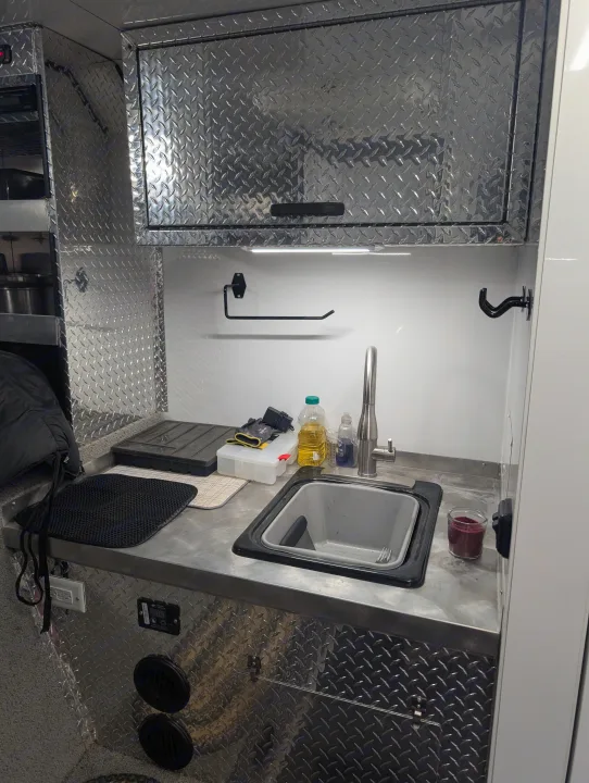This rv rental kitchen features a durable diamond plate counter and cabinets, with a sink, lights, and storage.. Trails West RPM Toy Hauler 2023
