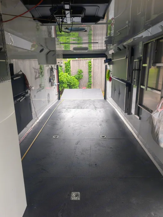 Spacious RV rental interior with reflective walls, black floor, and open ramp. View through back door to lush fence.. Trails West RPM Toy Hauler 2023