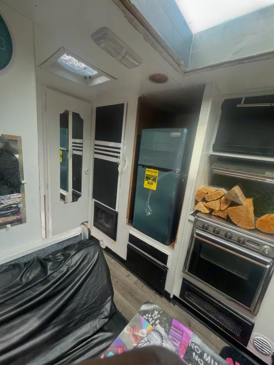 Interior of an RV rental showing a compact refrigerator, stove with firewood, and black faux leather seating.. Skyline Century 1985