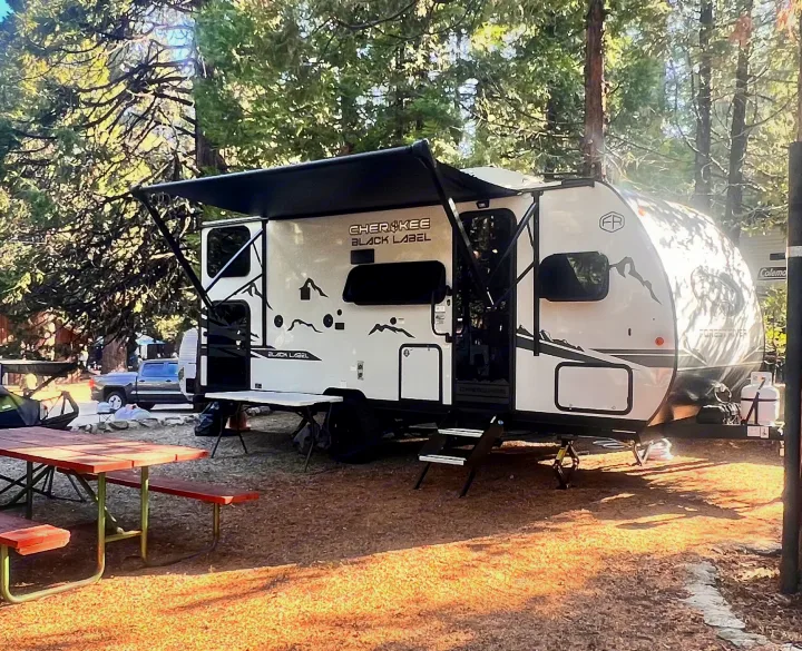 Cherokee Black Label rv rental parked in a shaded forest campsite with a picnic table and truck nearby. Perfect for outdoor escapes.. Cherokee WOLF PUP BLACK LABEL 17JWBL 2025