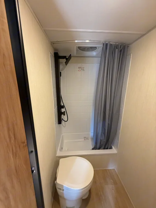 Compact RV rental bathroom with a white toilet, tub/shower, grey curtain, and ceiling vent fan.. Cherokee WOLF PUP BLACK LABEL 17JWBL 2025