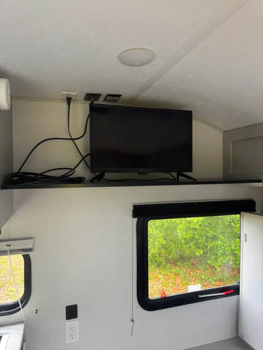 RV rental interior: a black TV on a shelf, power outlets, and two windows with blinds revealing an outside green view.. Forest River Salem FSX PLATINUM 2021