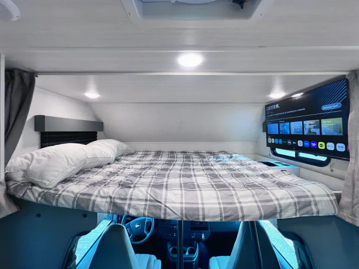 Cozy overhead bunk bed in an RV rental, featuring a plaid comforter and a TV, providing comfortable sleeping and entertainment.. East to West Entrada 2025