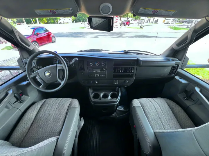 Inside an RV rental, this spacious van cabin features gray seats, dashboard controls, steering wheel, and a view of parked vehicles outside.. East to West Entrada 2025