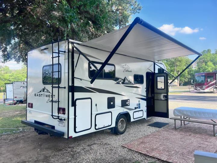 This white East West Entrada RV rental with an extended awning is set up at a shaded campsite, next to a picnic table.. East to West Entrada 2025