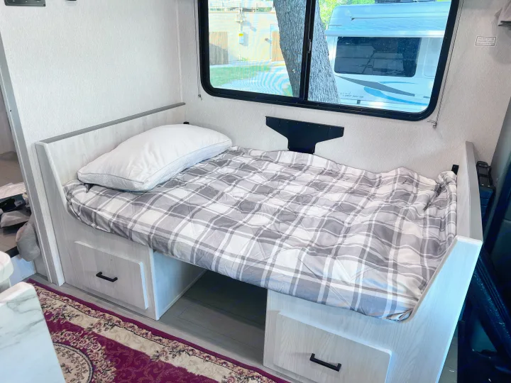 Cozy RV rental bed with plaid comforter, pillow, and under-bed storage drawers. Window view outside.. East to West Entrada 2025