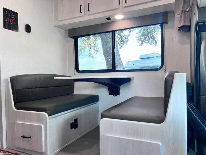 Modern RV rental dinette with light wood benches, dark cushions, a wall-mounted table, window view, and a control panel.. East to West Entrada 2025