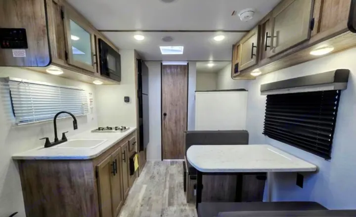 View of an rv rental's kitchen and dining area. Features wood-grain cabinets, sink, stove, microwave, and a dining table with bench seats.. Gulf Stream Kingsport Lite 199DD 2025