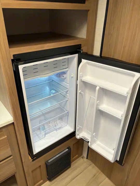 An open, empty, and clean mini-fridge in an RV rental. Its bright interior shows shelves, a freezer, and door storage, ready for your items.. Keystone RV Coleman 2025