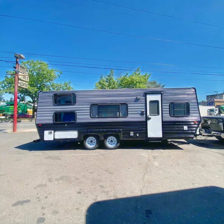 Black and grey striped RV rental (travel trailer) parked on asphalt, hitched to a pickup truck under a clear sky.. Skyline Century 1985