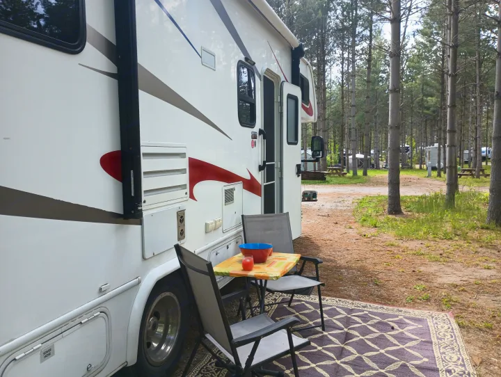 Experience camping freedom with an rv rental. RV parked in a shaded forest campground with outdoor chairs and table.. Forest River Sunseeker 2018