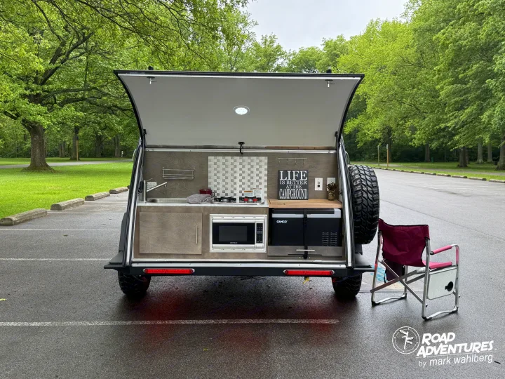 Teardrop RV rental showcasing its open outdoor kitchen: sink, stove, microwave, fridge, and 'Life is Better' sign. Perfect for camping!. nuCamp T@G XL 2025