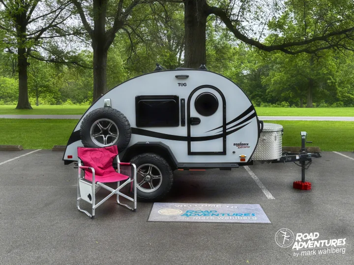 A sleek T@G teardrop RV rental awaits road adventures, set with a pink chair and welcome mat in a scenic outdoor spot.. nuCamp T@G XL 2025