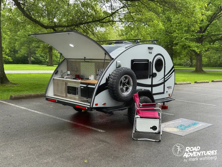This T@G teardrop camper RV rental's outdoor kitchen is open for road adventures. A chair sits ready in a scenic park.. nuCamp T@G XL 2025