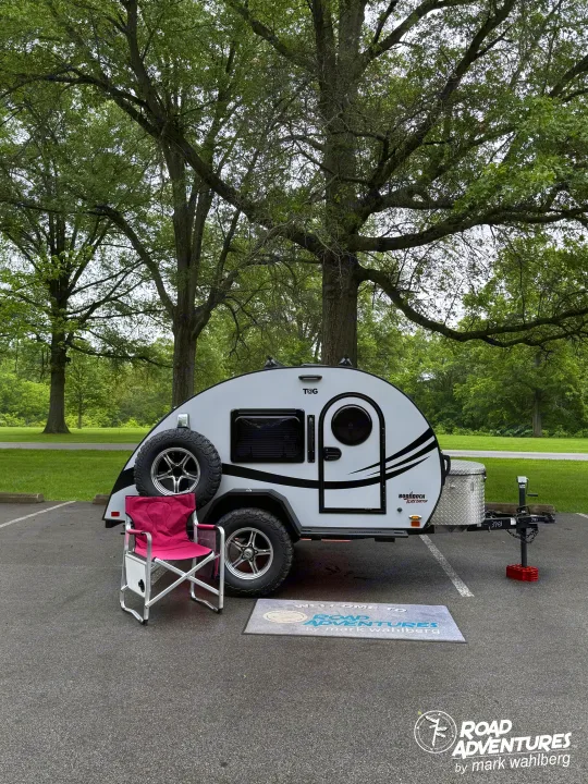 This compact T@G teardrop rv rental is ready for camping, featuring a pink chair and mat in a tree-lined parking spot.. nuCamp T@G XL 2025