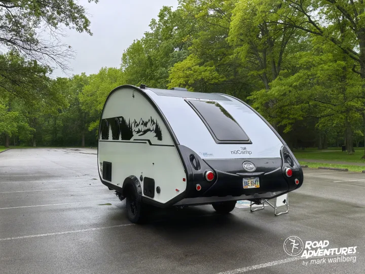This nuCamp teardrop travel trailer, an excellent RV rental option, gleams white & black on a wet lot surrounded by green trees.. nuCamp T@B 2025