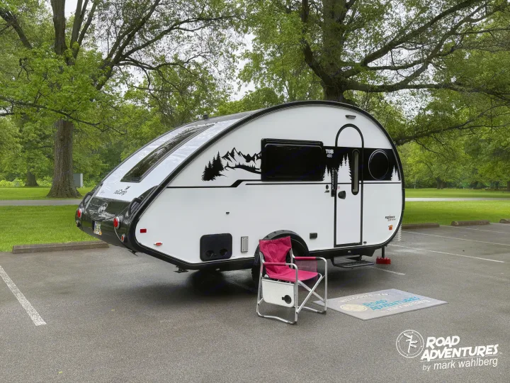 White T@B 400 teardrop rv rental camper with a pink folding chair and "Road Adventures" mat, parked in a green park.. nuCamp T@B 2025