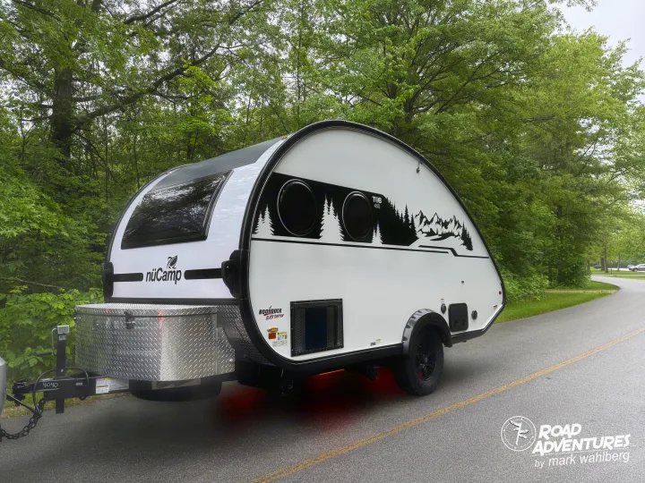 This nuCamp T@B 400 teardrop camper, ideal for an rv rental, is parked on a road surrounded by lush trees.. nuCamp T@B 2025