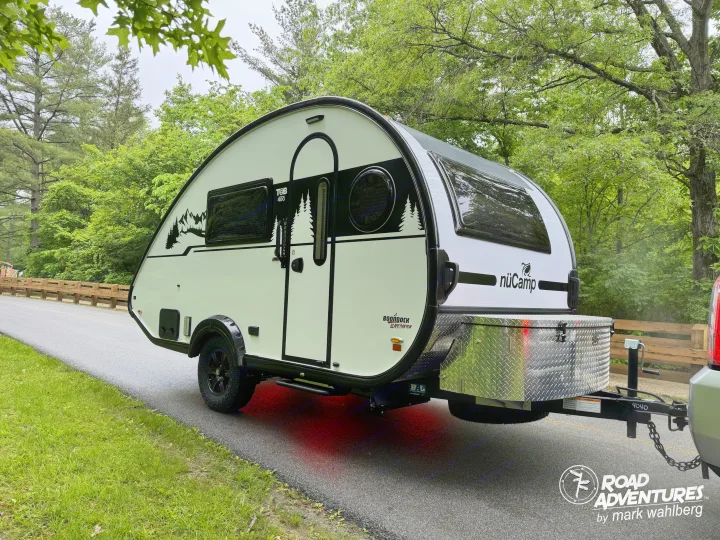 This white nuCamp T@B 400 teardrop travel trailer, an ideal RV rental for outdoor adventures, is parked on a road with green trees.. nuCamp T@B 2025