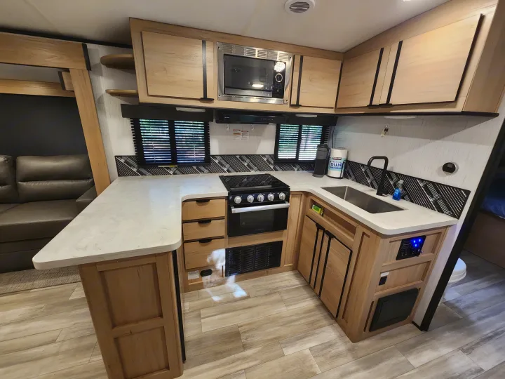 Modern rv rental kitchen featuring light wood cabinetry, countertop, sink, stove, microwave, and patterned backsplash. Ideal for travel.. Cruiser RV Radiance 2022