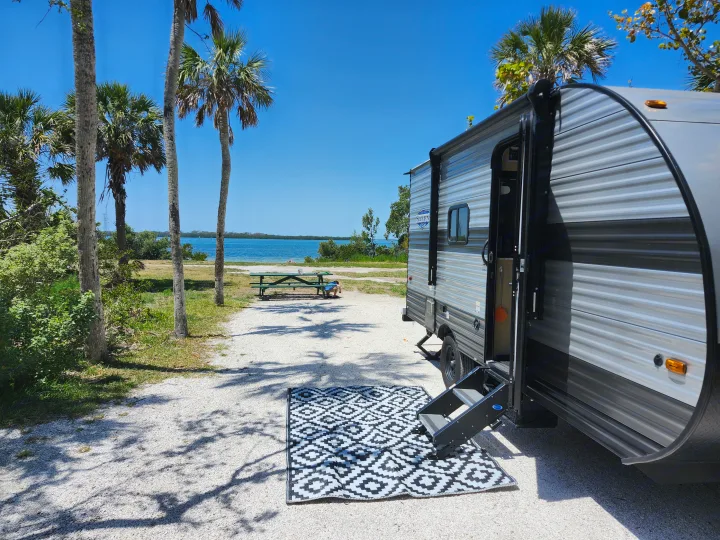 RV rental parked at a serene, sunny waterfront campsite. Features an outdoor rug, palm trees, and a picnic table by blue water.. Forest River Salem FSX 2023