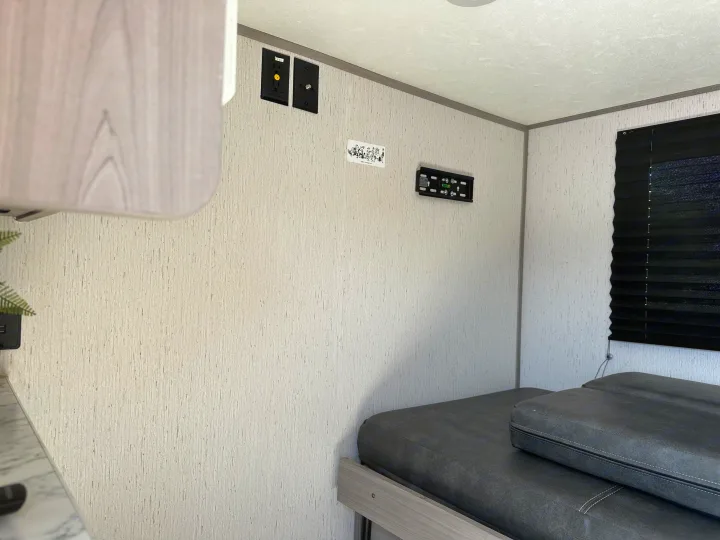 Inside an RV rental, featuring a dark grey bed, TV mount, electrical outlets, and a black window blind.. Dutchmen Coleman Rubicon 2023