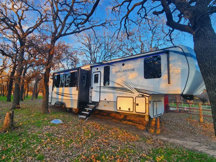 This large white Avalanche fifth-wheel RV is an ideal RV rental, parked amidst bare trees and fallen leaves under a blue sky.. Keystone RV Avalanche 2023