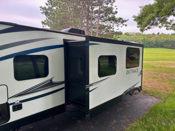 White Outback travel trailer RV rental with extended slide-out, parked outdoors by a field with trees.. Keystone RV Outback Ultra-Lite 2017