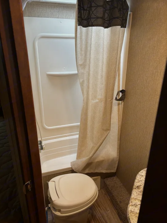 RV rental bathroom with white compact shower, built-in shelves, toilet, and a beige patterned shower curtain.. Keystone RV Outback Ultra-Lite 2017
