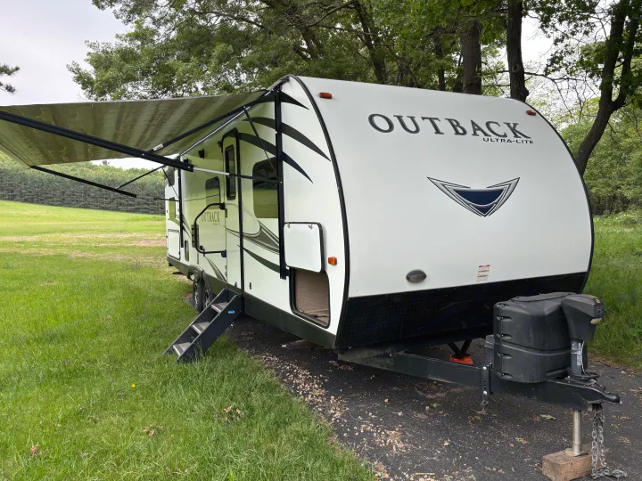 A white Outback Ultra-Lite travel trailer, an ideal RV rental, with an extended awning in a green, natural setting.. Keystone RV Outback Ultra-Lite 2017