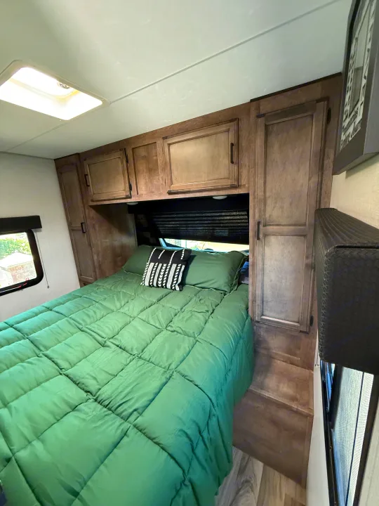 Cozy RV rental bedroom featuring a bed with a green comforter, decorative pillow, built-in cabinetry, and a window.. Sport Trek 270 VBH 2021