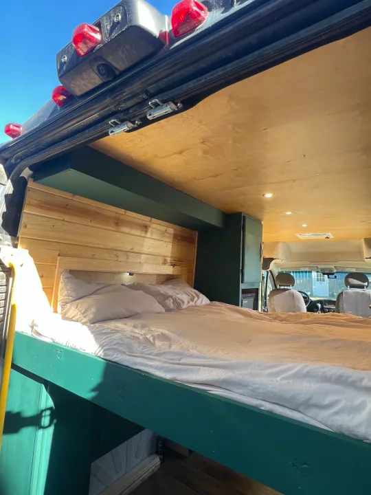 Bright RV rental interior: cozy bed with wood headboard, green frame, wood ceiling, and front cabin view.. Dodge ProMaster 2500 2018