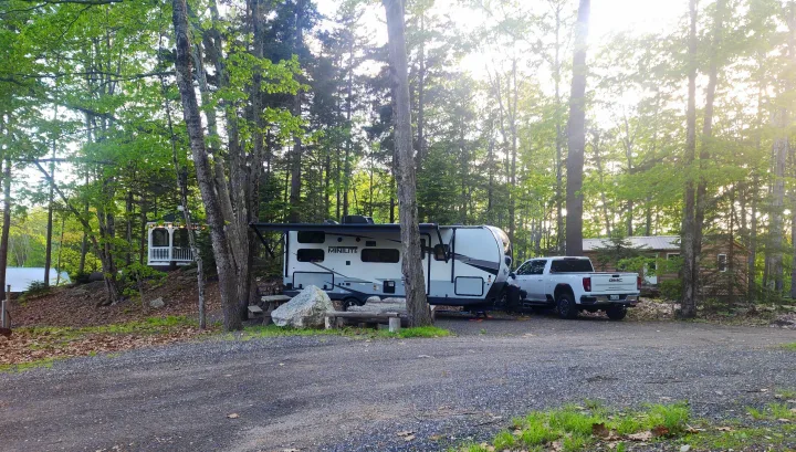 An RV rental travel trailer is parked with a truck at a wooded campsite, featuring a picnic area and gravel ground.. Forest River Rockwood Mini Lite 2023