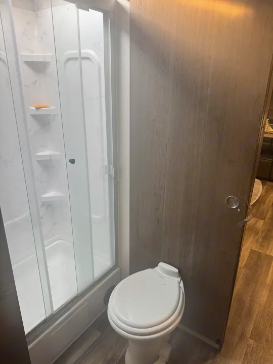 RV rental bathroom with a clear glass shower door, soap on a shelf, white toilet, and wood-paneled door.. Keystone RV Cougar 2022