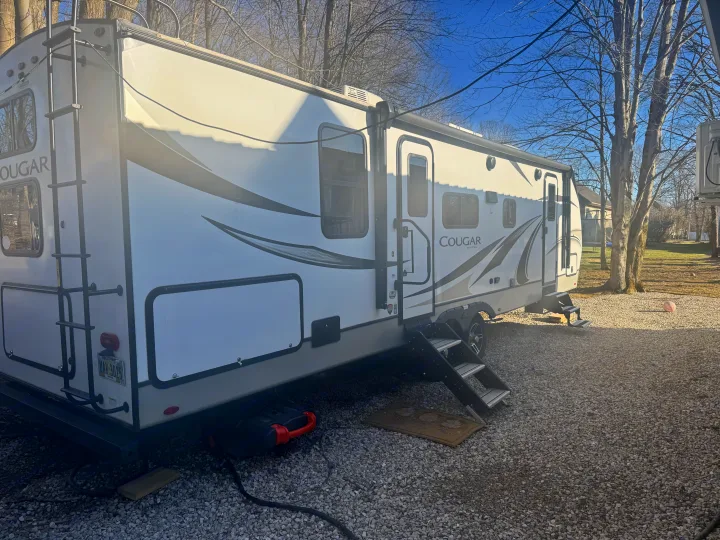 White Cougar RV rental travel trailer on a gravel site with steps, visible ladder, and power hookup.. Keystone RV Cougar 2022